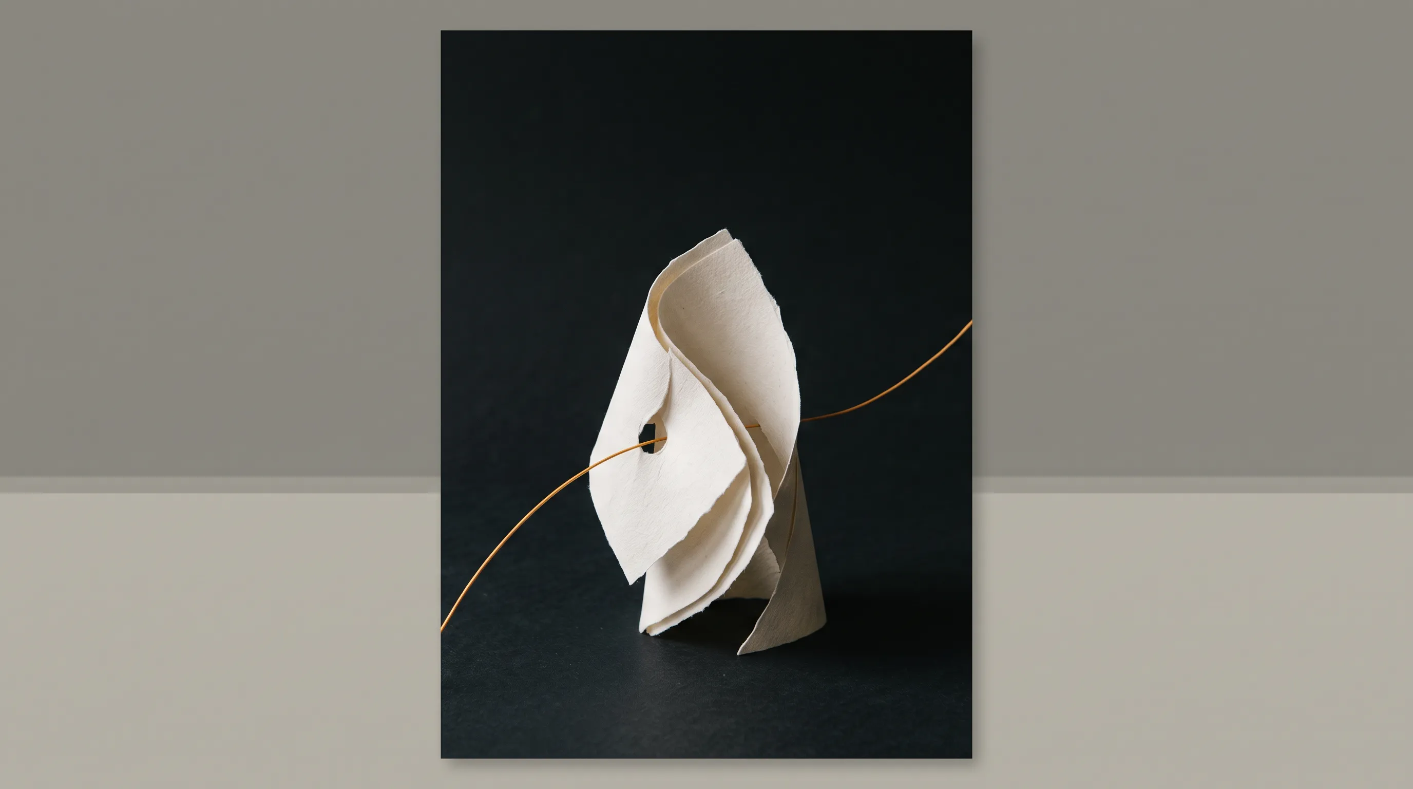 A folded warm off-white paper form on a near-black surface, crossed by a fine amber line under soft directional light.